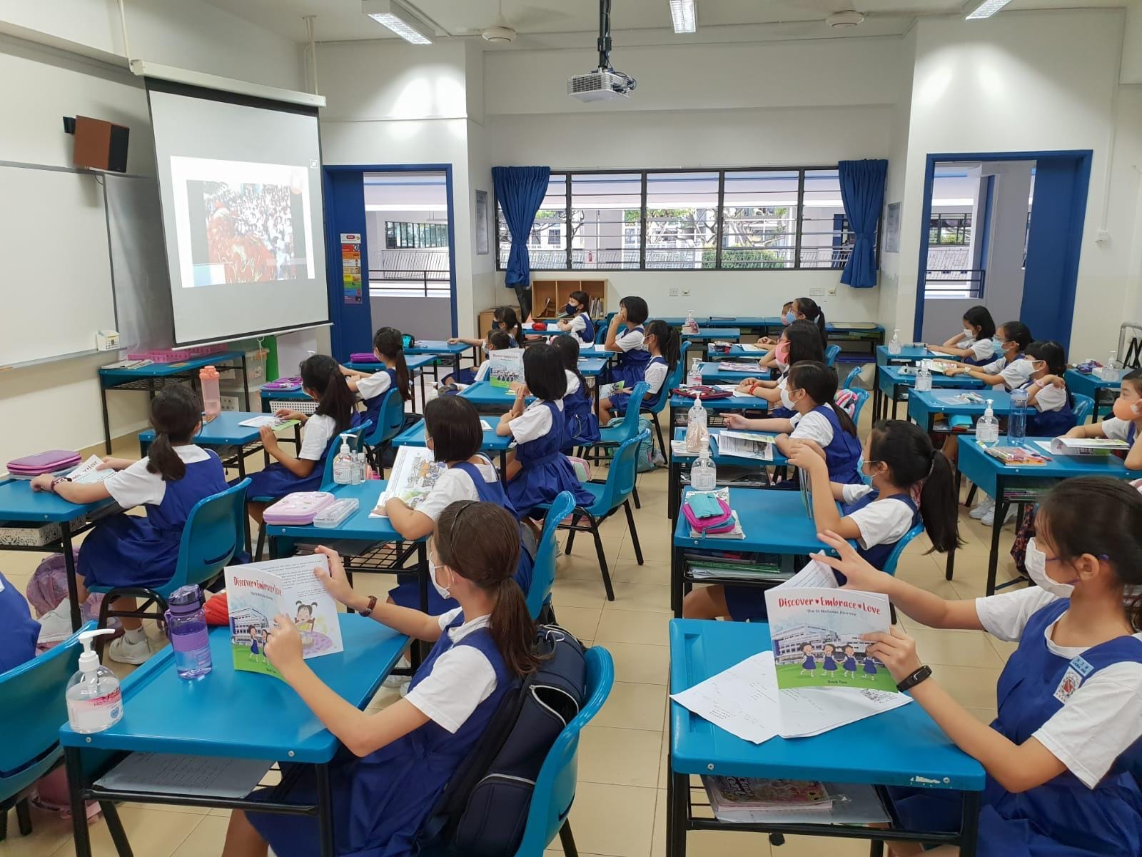 Students in blue uniforms sit at blue desks in a classroom, holding booklets and facing a projected screen.